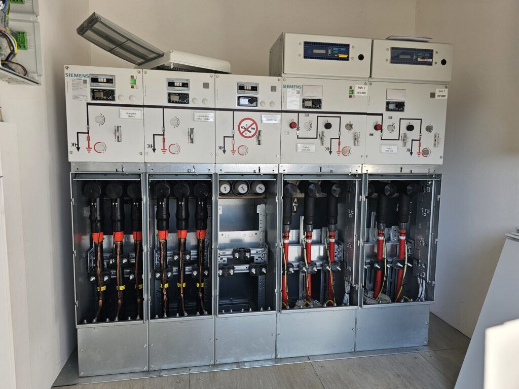 Mittelspannungs‑Schaltanlage von Siemens mit mehreren Schaltfeldern, oben Bedienelemente und Anzeigen, unten Kabelabgänge mit roten und schwarzen Leitungen.