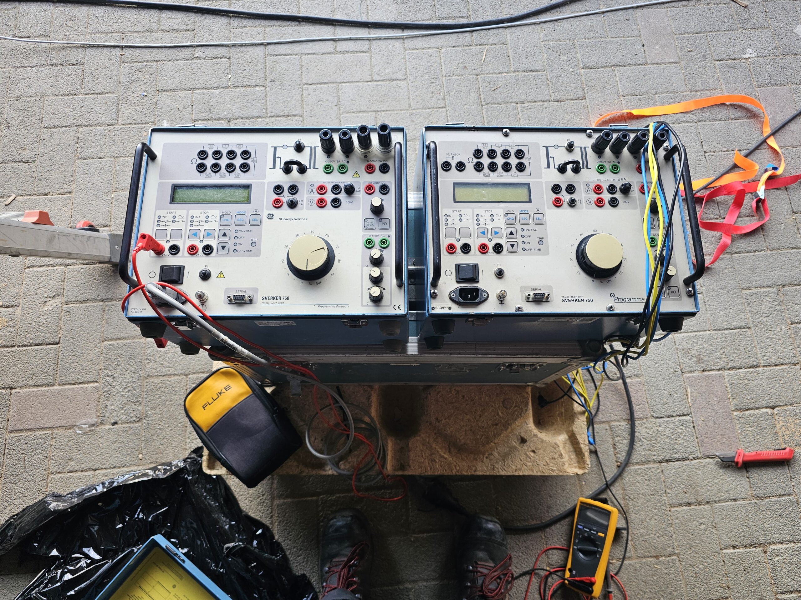 Zwei SVERKER‑Prüfgeräte zur Sekundärprüfung von Schutzrelais auf Holzpalette aufgestellt, mehrere Messleitungen und Multimeter angeschlossen.