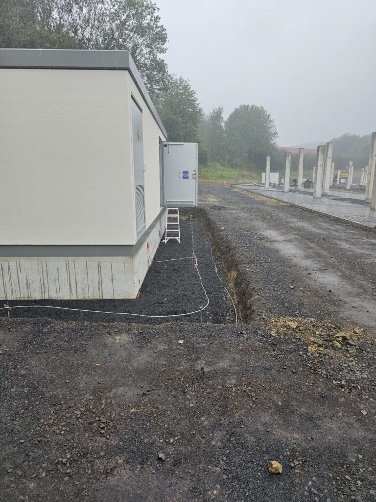 Stationsgebäude mit offenem Graben für Erdungsanlage auf Baustelle bei bewölktem Wetter.