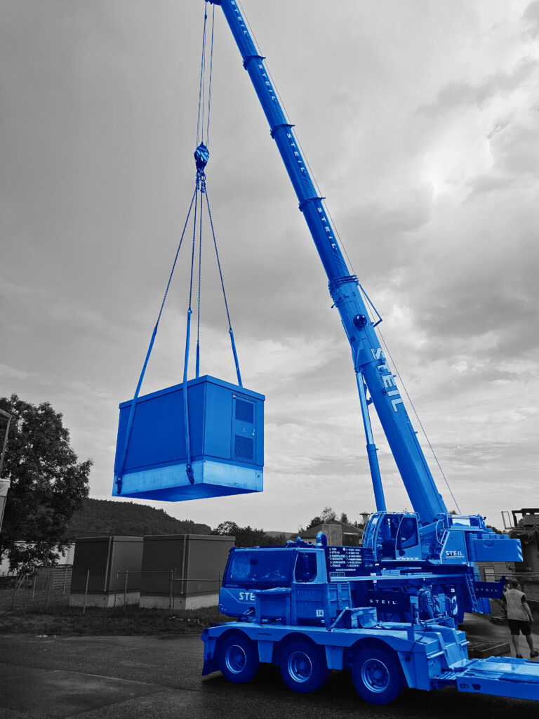 Blauer Autokran hebt blauen Container über Asphaltfläche bei bewölktem Himmel.