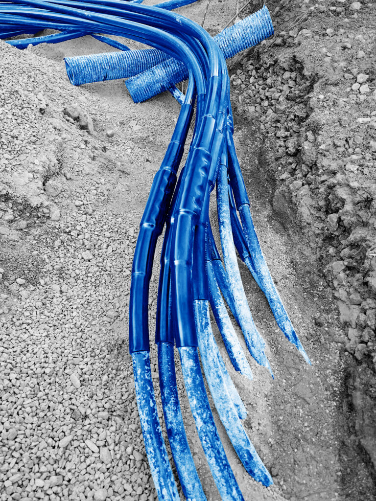 Mehrere blaue Kabelschutzrohre liegen in offenem Graben auf Schotter.