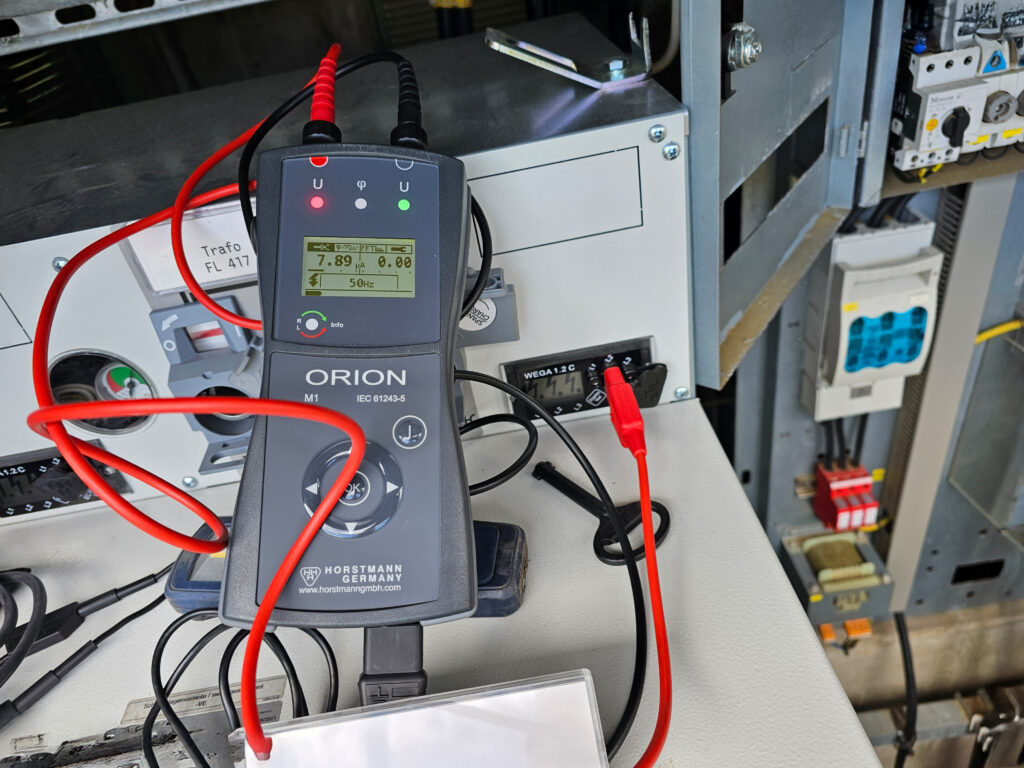 Orion‑Messgerät mit roten Messleitungen an Trafoanlage zur Spannungsprüfung angeschlossen.