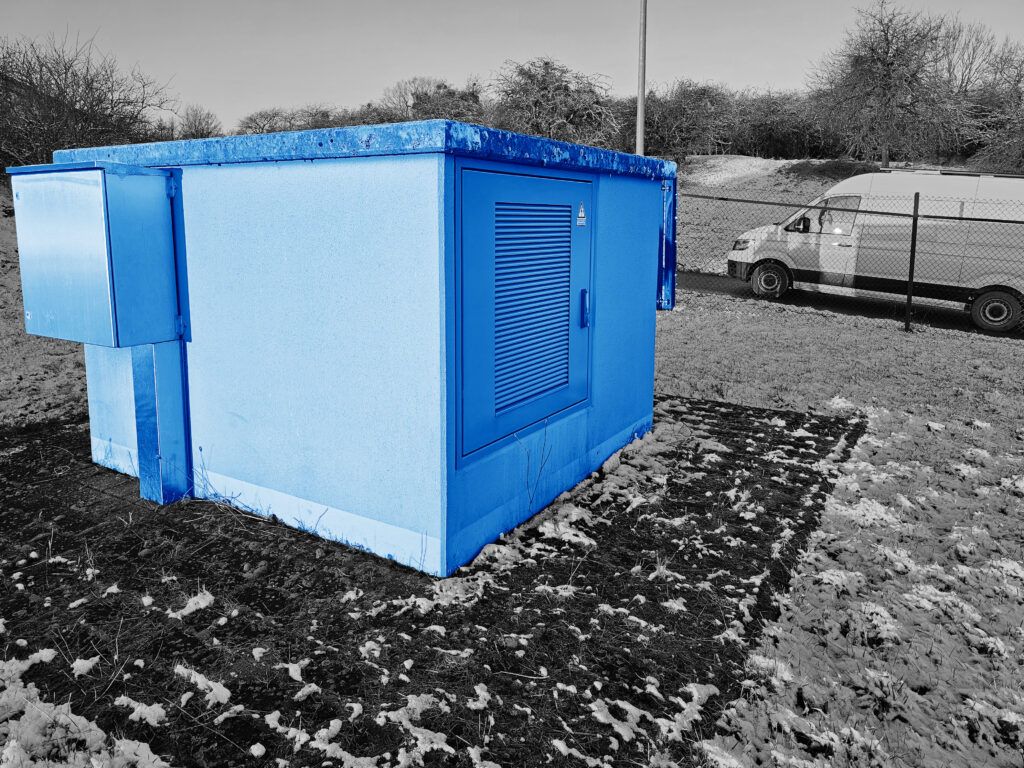 Blaue Trafostation auf freier Fläche und seitlichem Anschlusskasten, im Hintergrund Servicefahrzeug.