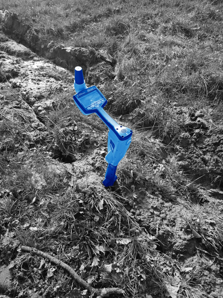 Blaues Vivax‑Ortungsgerät mit Display und Sensorstab auf Gras und Erde.